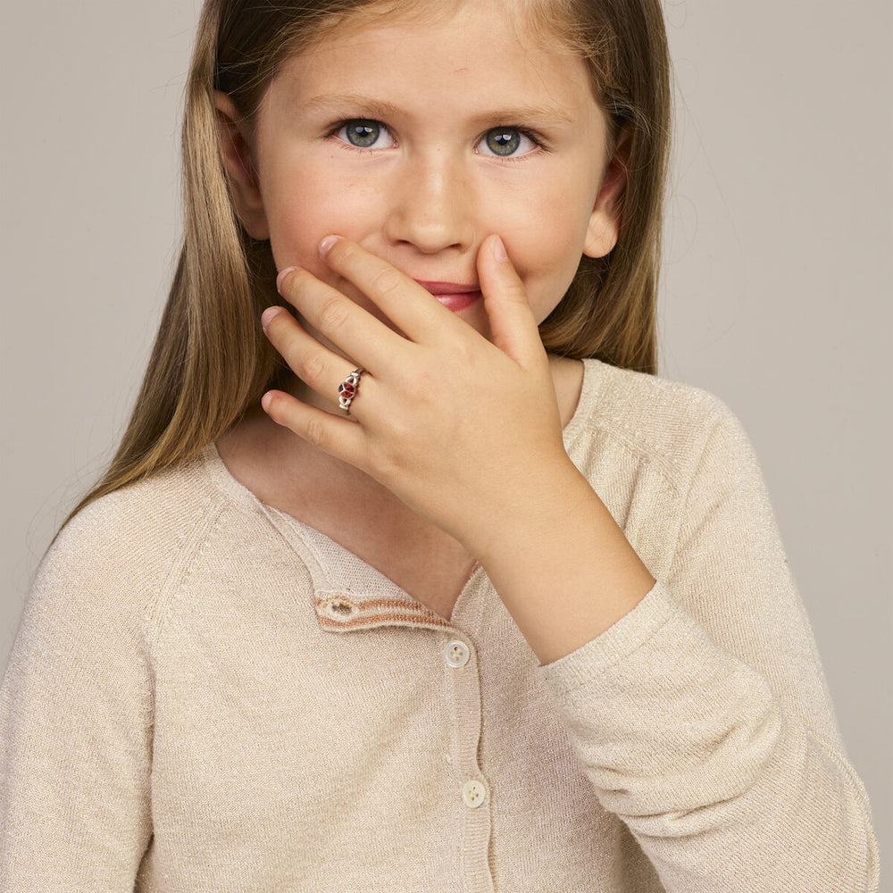 Een jong meisje met lang lichtbruin haar en blauwe ogen, dat een beige vestje draagt, glimlacht zachtjes en bedekt haar mond met één hand, terwijl ze een Van der Kooij Collectie ring lieveheersbeestje zilver wit om haar vinger laat zien.