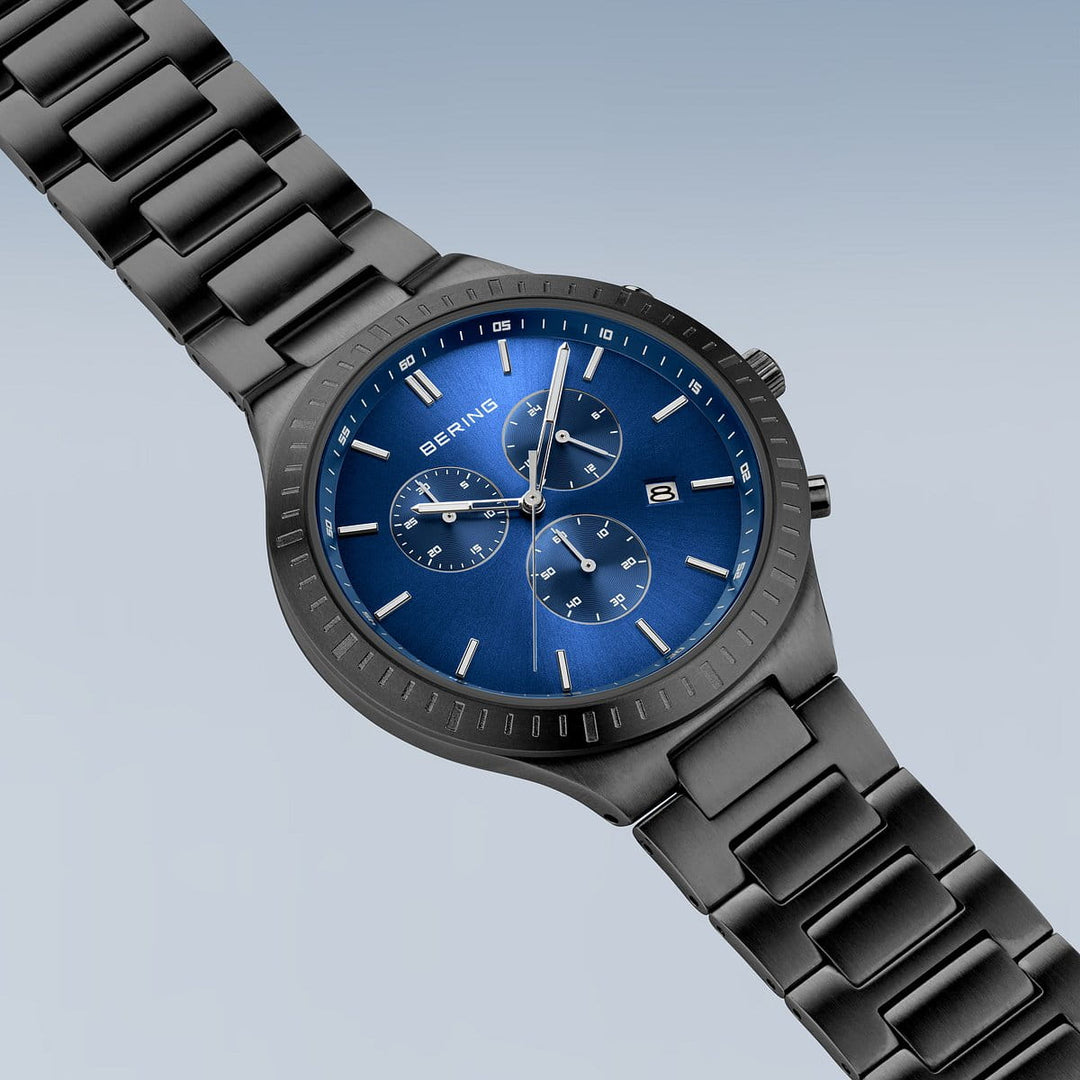 De Bering Herenhorloge 11743-727 heeft een blauwe wijzerplaat met drie sub-wijzerplaten, datumweergave, zilveren uurmarkeringen en een metalen band. Gemaakt van titanium, heeft het een strak design tegen een licht gradiënt achtergrond.