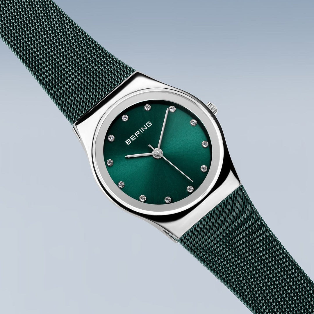 Het Bering dameshorloge, model 12927-808, heeft een groene wijzerplaat met zilveren uurmarkeringen in een minimalistisch design. Het omvat een bijpassend groen Milanees bandje en gepolijste zilveren lunette op een gradiëntblauwe achtergrond, met de merknaam Bering elegant weergegeven.