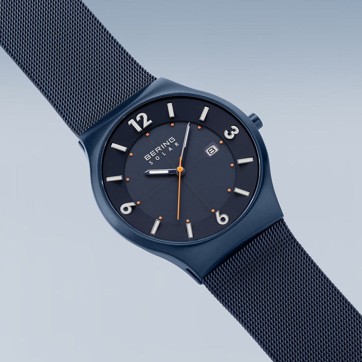 Het Bering-model 14440-393 wordt schuin weergegeven en heeft een blauw zonne-uurwerk en een mesh-band. Het horloge heeft een zwarte wijzerplaat met witte markeringen, een secondewijzer met oranje punt en een klein datumvenster onder duurzaam saffierglas op een achtergrond die van licht naar donkerblauw verloopt.