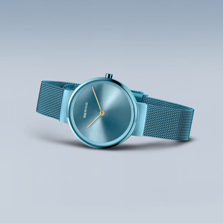 De Bering 14531-388 is een strak blauw dameshorloge, voorzien van een mesh band en minimalistische wijzerplaat met zilveren markeringen en wijzers, beschermd door saffierglas. Het wordt elegant weergegeven tegen een lichtblauwe achtergrond met kleurverloop.