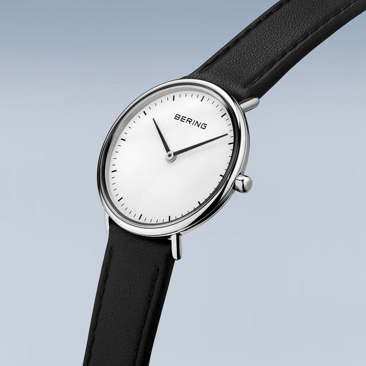 De Bering 15729-404 is een minimalistisch dameshorloge met een witte wijzerplaat, geen cijfers, dunne zwarte uur- en minutenwijzers, een strakke zilveren kast beschermd door saffierglas en een zwarte leren band op een lichtblauwe achtergrond.