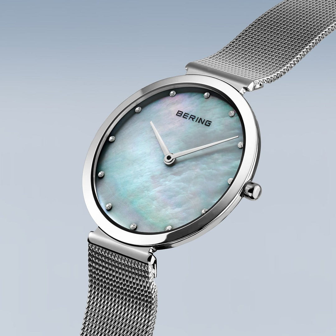 De Bering 18132-004 is een strak dameshorloge met een zilveren meshband en ronde wijzerplaat. Het heeft een parelmoeren wijzerplaat met minimaal design, dunne zilveren wijzers, kleine zilveren uurmarkeringen en saffierglas bescherming. Het merk BERING is prominent weergegeven bovenaan de wijzerplaat.