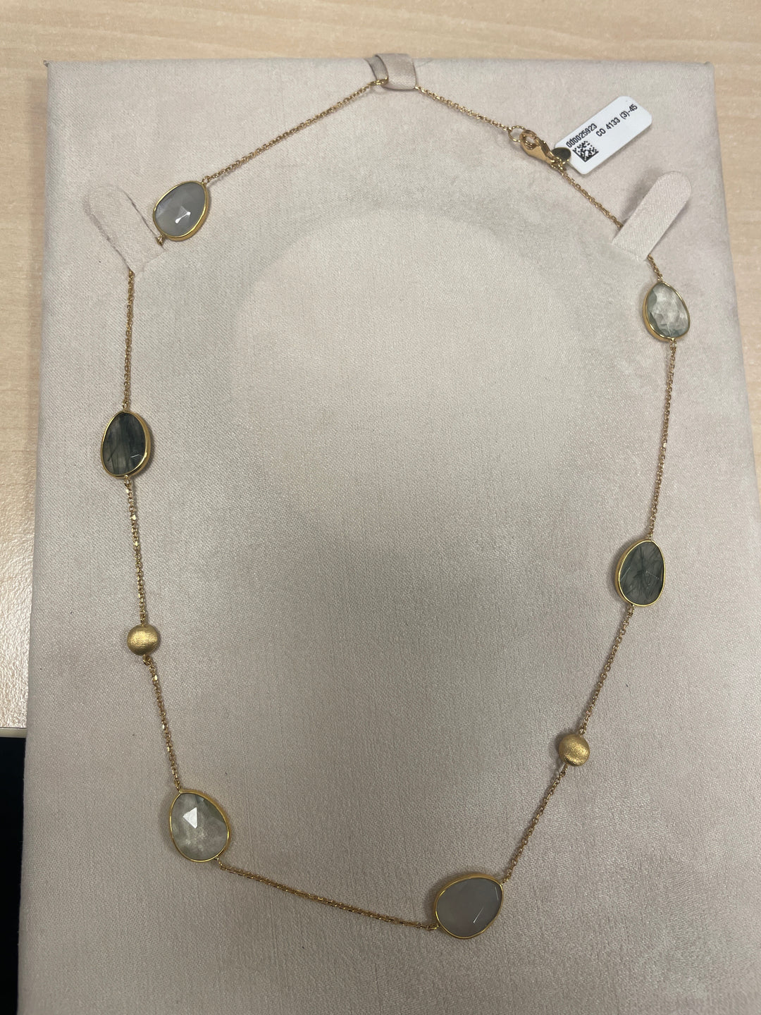 De Italo Bottene CO 4133 (3)-45 ketting is een delicaat, goudkleurig sieraad met ovale stenen in gedempte grijs- en groentinten, afgewisseld met kleine gouden kraaltjes, elegant tentoongesteld op een beige gewatteerd oppervlak.