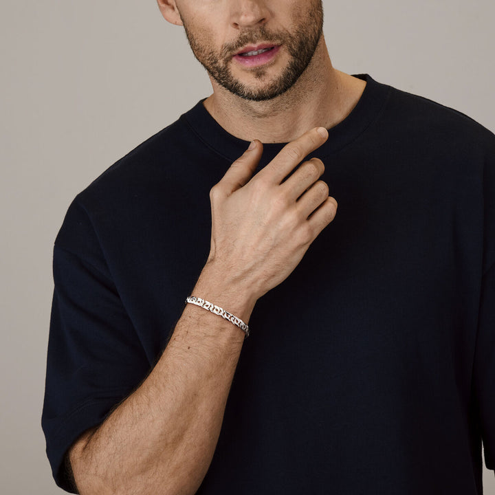 Een man met een bijgeknipte baard en een donkerblauw t-shirt poseert met zijn hand bij zijn gezicht en toont de armband van 6,5 mm zilverwit van Van der Kooij Collectie om zijn pols.