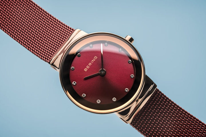 Het Bering Dameshorloge, model 10126-363, heeft een dieprode wijzerplaat met kristallen markeringen en een roségouden kast. Het heeft een Quartz uurwerk en een bijpassende rode meshband, allemaal tegen een lichtblauwe achtergrond.