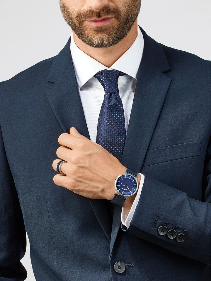 Een man in een marineblauw pak trekt zijn blauwe stropdas recht en toont een strak Bering-horloge (model 15439-307) met saffierglas op zijn pols. Zijn witte overhemd is elegant gecombineerd met het zwarte jasje met knopen en zijn rechterhand is versierd met een ring tegen de lichte achtergrond.