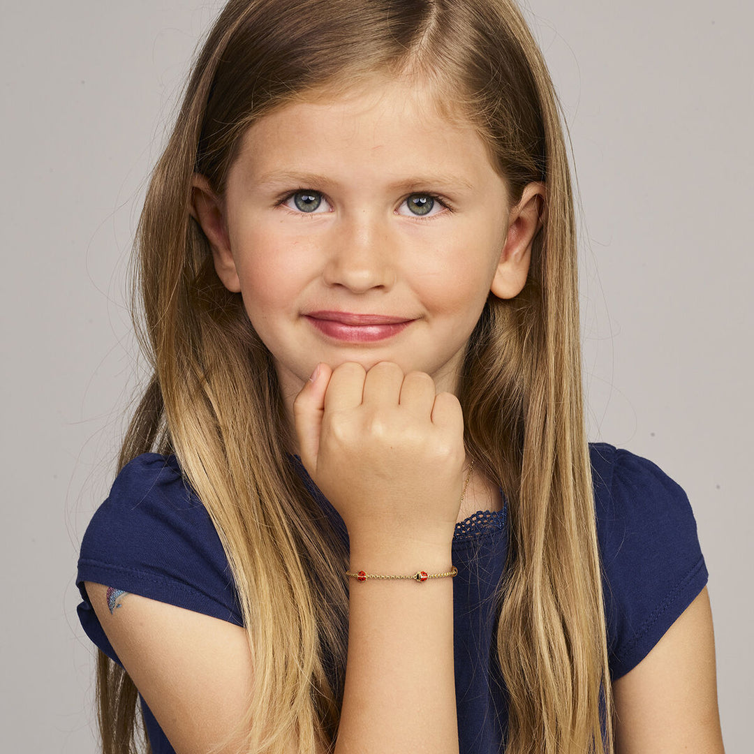Een jong meisje met lang blond haar en een marineblauw shirt glimlacht naar de camera, haar hand onder haar kin, met om haar pols de Van der Kooij Collectie armband lieveheersbeestje 11-13 cm in 14K geelgoud.