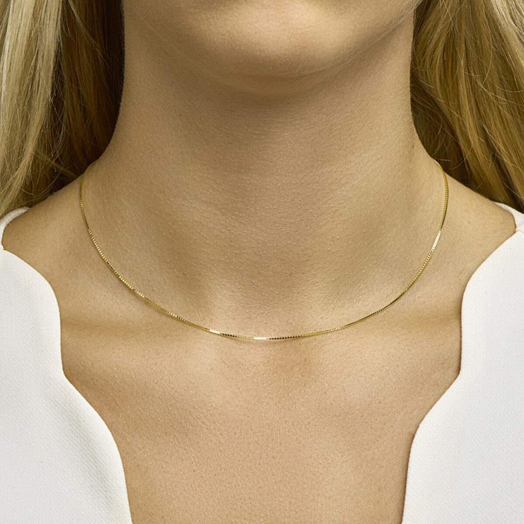 Een vrouw draagt het delicate Collier Venetiaans 0,8 mm 14K geelgoud uit de Van der Kooij Collectie, een dun gouden collier met een Venetiaanse schakel, dat haar blote nek siert boven een wit topje met V-hals.
