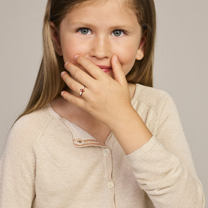 Een jong meisje met lang lichtbruin haar en blauwe ogen draagt een beige vest en bedekt haar mond. Ze toont een Van der Kooij Collectie ring lieveheersbeestje zilver wit. Ze kijkt naar de camera met een verlegen glimlach.
