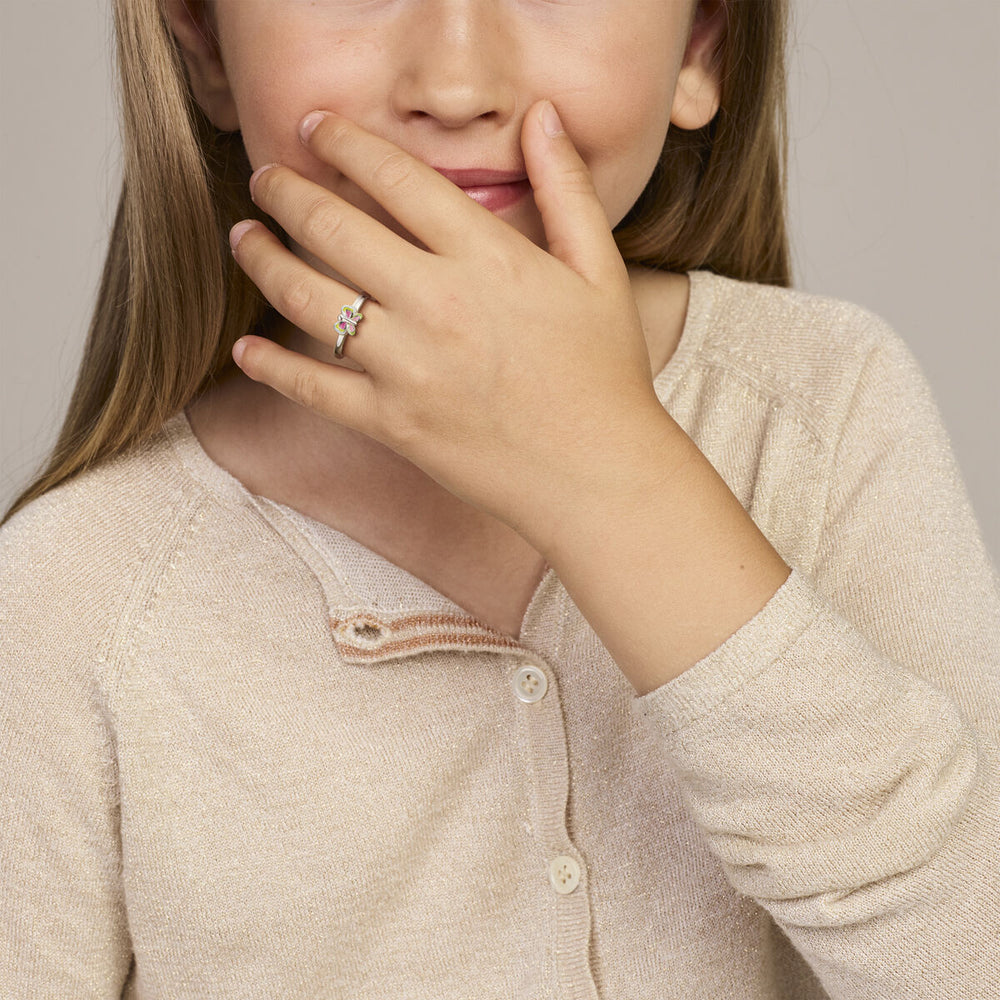 Een jong meisje met lang haar, gekleed in een beige vest, bedekt haar mond, waardoor een Van der Kooij Collectie ring vlinder zilver gerhodineerd om haar vinger zichtbaar wordt. Ze lacht zachtjes.