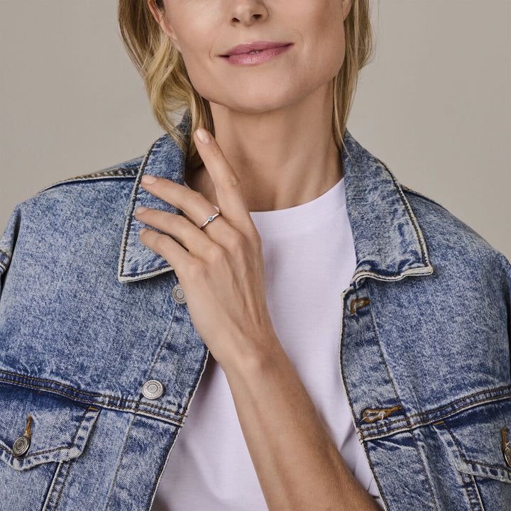 Een vrouw in een licht spijkerjasje en wit t-shirt showt de Van der Kooij Collectie ring met geboortesteen blauw topaas december zilver gerhodineerd aan haar linkerhand, zachtjes tegen haar kraag tegen een neutrale achtergrond.