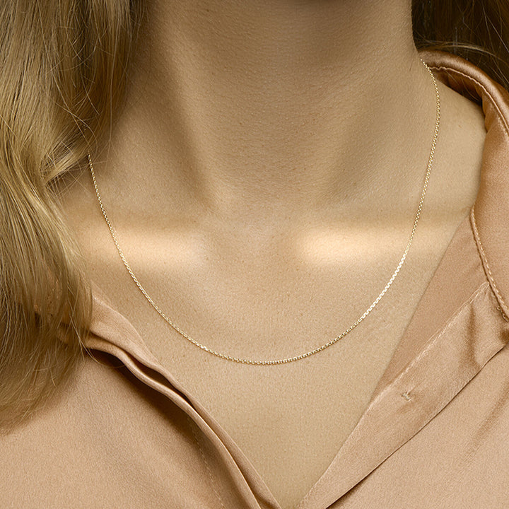 Een close-up toont een persoon die de Van der Kooij Collectie collier anker gediamanteerd 1,1 mm 41 + 4 cm 3 micron zilver verguld (geel) ketting draagt over een lichtbruine satijnen blouse; alleen de hals, het sleutelbeen en een deel van de blouse zijn zichtbaar.