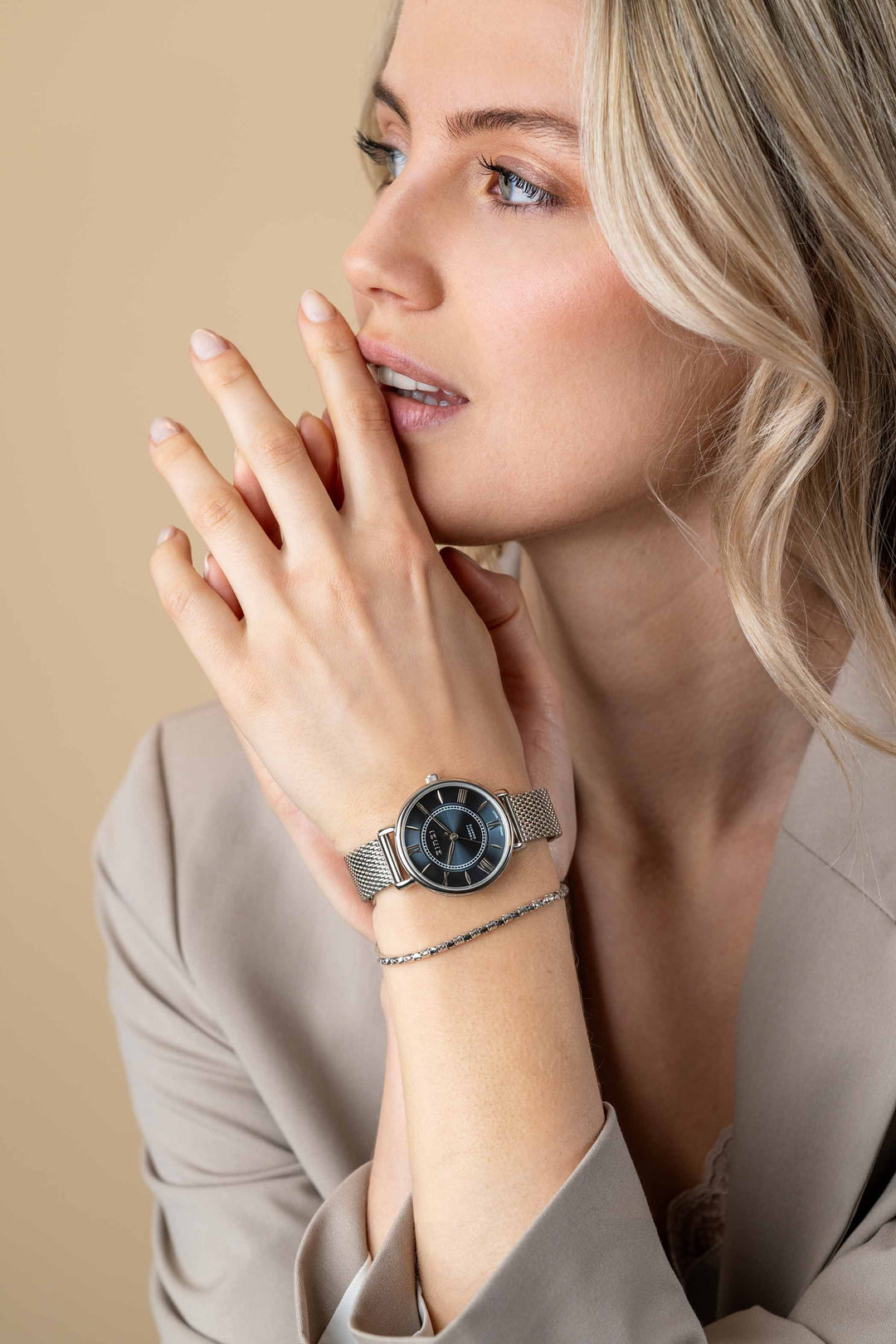 Een vrouw met blond haar kijkt peinzend voor zich uit en raakt haar lippen aan. Ze draagt een beige blazer, het Zinzi zilverkleurige Horloge Ziw2053 met zwarte wijzerplaat van Zinzi en een delicate zilveren armband.