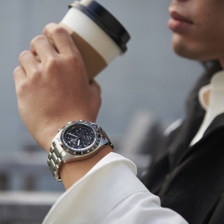 Het individu, getooid met een Seiko Prospex herenhorloge SSC819P1 met een roestvrijstalen kast, houdt een papieren koffiekopje vast met een zwart deksel. Gekleed in een wit overhemd en een donker jasje, is hun gezicht gedeeltelijk zichtbaar tegen de wazige achtergrond.
