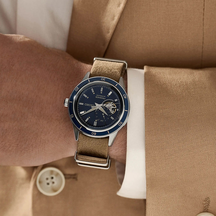 Een persoon in een bruin pak en wit shirt is te zien met de borst naar beneden, met het Seiko Presage heren horloge SSA453J1. Dit automatische horloge heeft een blauwe wijzerplaat, bruine band, zichtbare tandwielen, Romeinse cijfermarkeringen en duurzaam Hardlex mineraalglas.