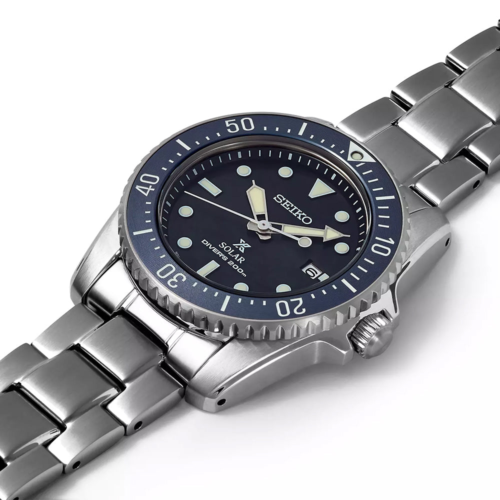 Het Seiko Prospex SNE569P1 Solar Divers 200m horloge heeft een roestvrijstalen kast en een zilveren metalen band. De blauwe wijzerplaat heeft lichtgevende markeringen, wijzers en een datumweergave op 3 uur, terwijl de lunette minutenindicatoren heeft voor nauwkeurige timing.
