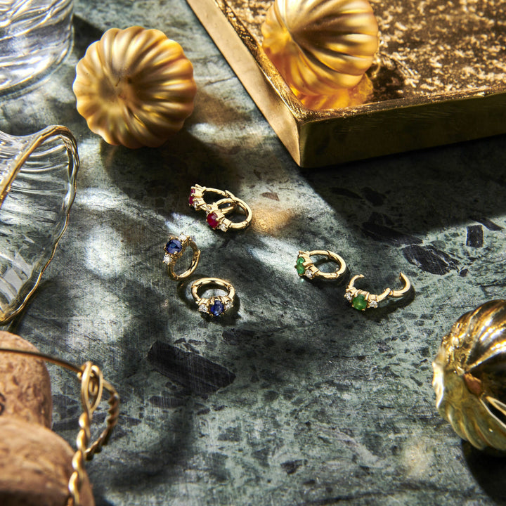 Van der Kooij Collectie klapoorringen saffier en diamant 0.10ct (2x0.05ct) h si, 14K geelgoud, zijn te zien op een groene, textuur ondergrond tussen en glazen decoraties, met zacht zonlicht dat lichte schaduwen werpt.