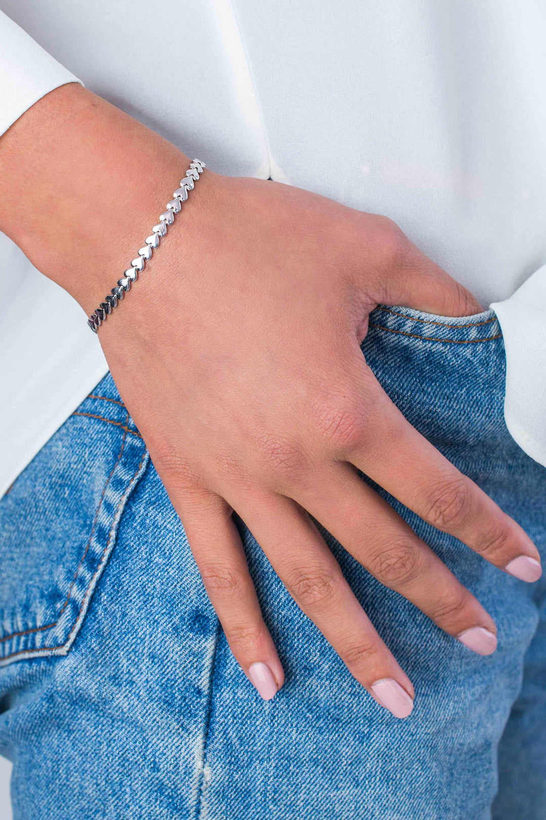 Een persoon die de Zinzi Armband Zilver Zia1798 van Zinzi draagt, legt zijn hand op de zak van een lichtblauwe spijkerbroek. Hun nagels zijn netjes gemanicuurd en ze dragen een witte top met lange mouwen.