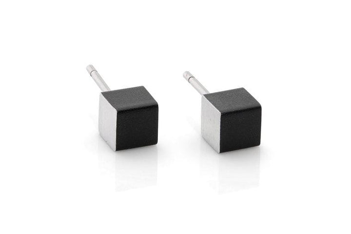 Twee kleine kubusvormige oorstekers met zwarte bovenkanten en zilveren steeltjes worden elegant tentoongesteld op een wit reflecterend oppervlak. De Oorsieraad Kubusjes Zwart O28Z van CLIC Creations zijn hypoallergeen en combineren comfort en stijl voor gevoelige oren.