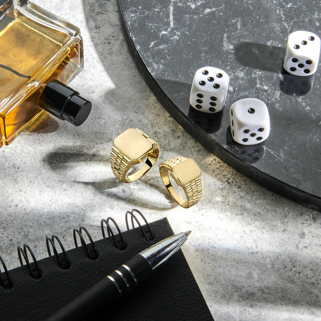 Een Van der Kooij Collectie graveerring gematteerd 14K geelgoud, een andere gouden ring, een glazen parfumflesje, drie witte dobbelstenen, een zwarte pen en een zwart spiraalvormig notitieboekje zijn gerangschikt op grijs marmer met zonlicht dat schaduwen werpt.