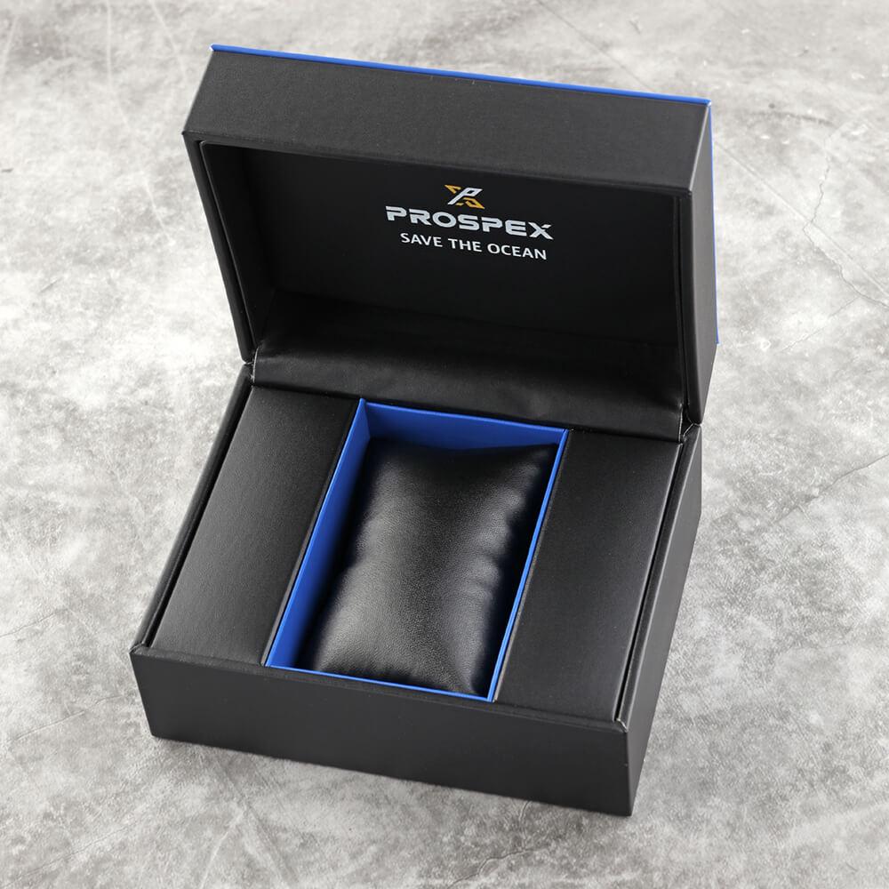 Een open Seiko Prospex SRPE35K1 doos met een blauwe binnenkant rust op een grijs oppervlak. Binnenin zit een zwarte kusseninzet, waarschijnlijk voor een horloge, en op het deksel staat PROSPEX SAVE THE OCEAN.