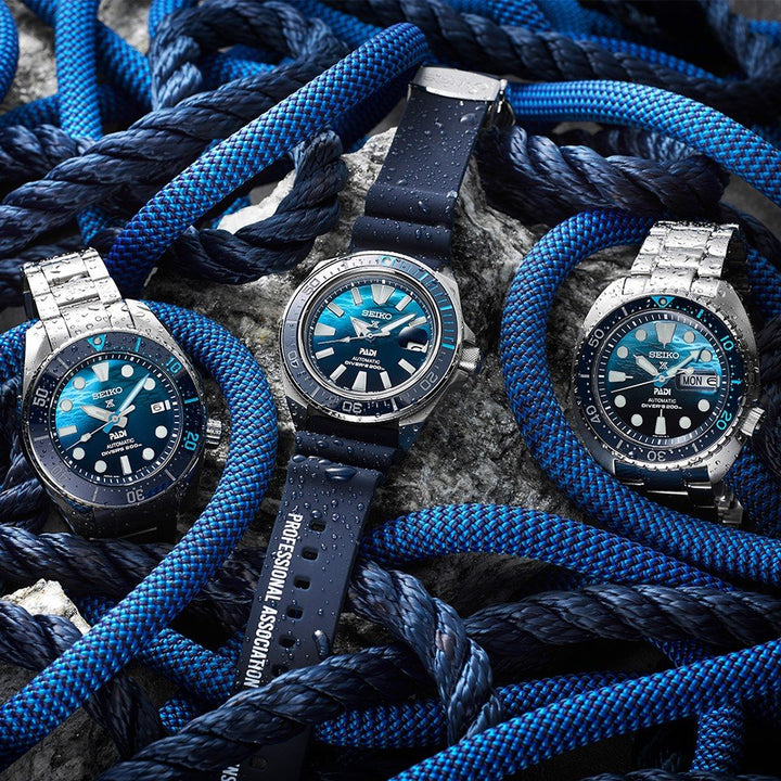 Drie Seiko Prospex duikhorloges, elk met blauwe wijzerplaten en roestvrijstalen kasten, hebben natte blauwe touwen. De linker- en rechterhorloges hebben metalen armbanden, terwijl het middelste horloge een rubberen band heeft. De gestructureerde achtergrond accentueert het nautische thema.