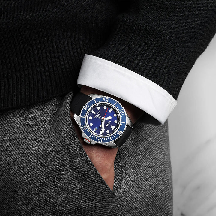 Een persoon draagt casual een stijlvol Seiko Prospex heren horloge SNE593P1 met een opvallende blauwe wijzerplaat en prominente markeringen. Het duikhorloge, met een zwarte band, complementeert hun donkere trui en witte overhemd terwijl hun hand moeiteloos in hun zak rust.