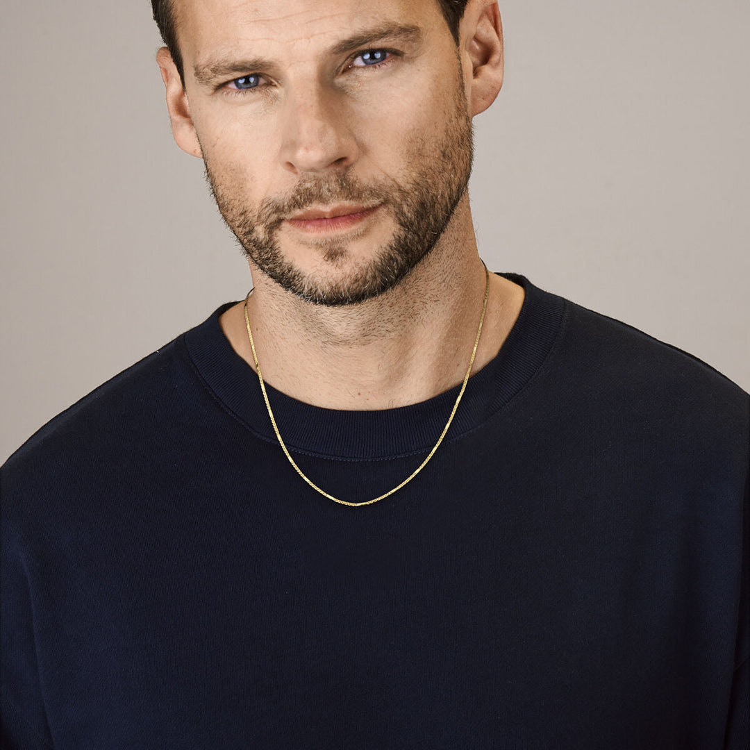 Een man met kort bruin haar, blauwe ogen en een getrimd baardje draagt een navy crewneck trui en een Van der Kooij Collectie 14K gouden ketting heren - collier valkenoog met tussenstuk 1,9 mm. Hij kijkt neutraal langs de camera tegen een effen achtergrond.
