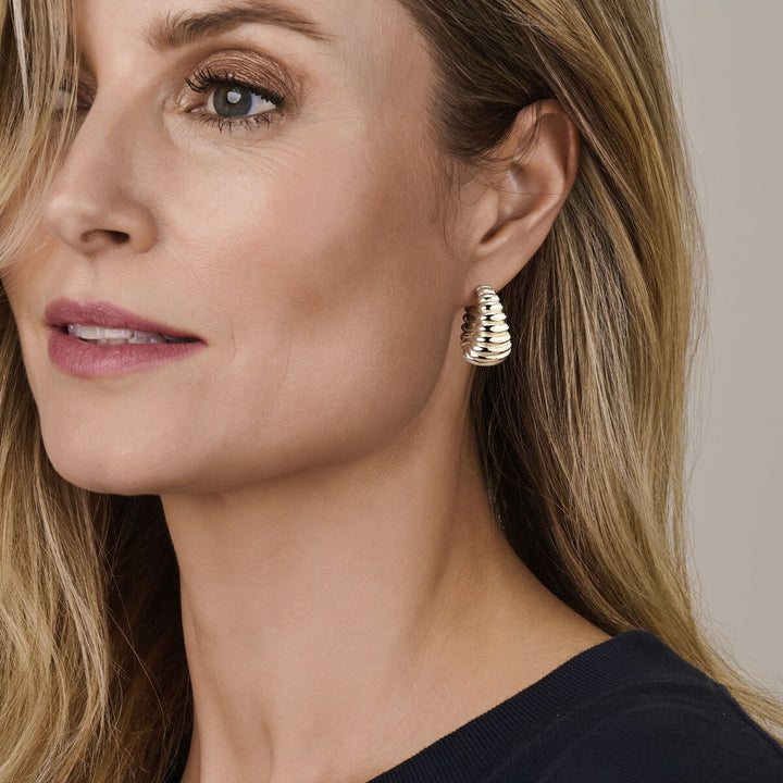 Een vrouw met lang blond haar in een marineblauwe top toont Van der Kooij Collectie oorringen 34 mm zilver gerhodineerd tegen een neutrale achtergrond, waardoor deze elegante damesaccessoires extra in de kijker staan.