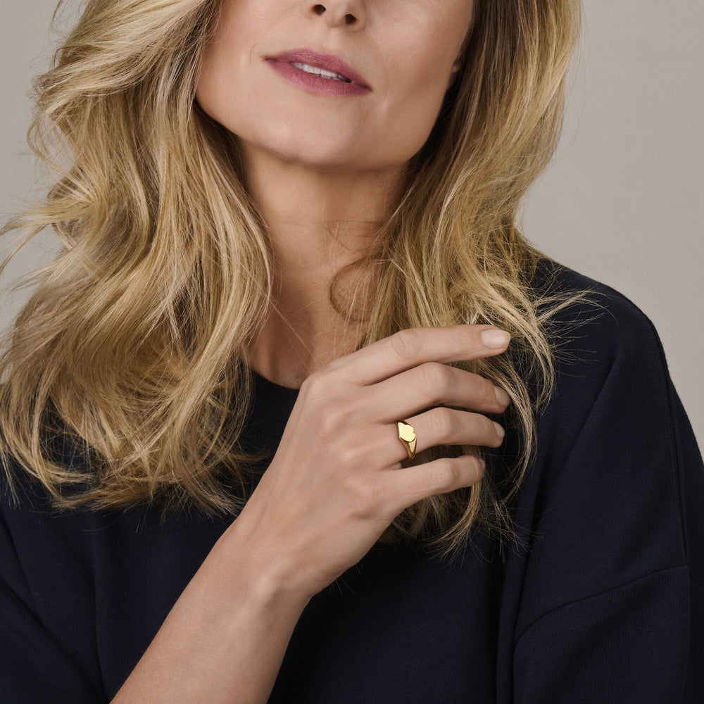Een vrouw met golvend blond haar en een donker topje toont een ring hartje 14K geelgoud van Van der Kooij Collectie aan haar hand, die ze bij haar gezicht houdt. Alleen de onderste helft van haar gezicht en haar hand zijn zichtbaar.