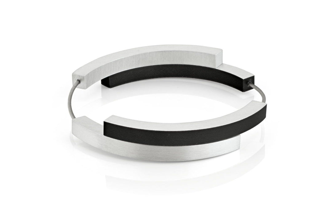 De Armband Vier bogen Zwart A32Z van CLIC Creations is een moderne, minimalistische bangle met afwisselende segmenten van zilver en zwart, verbonden door een dunne draad. Het vormt een halfopen cirkel voor een strakke, stijlvolle look. Handgemaakt in Nederland, het wordt weergegeven op een wit reflecterend oppervlak.