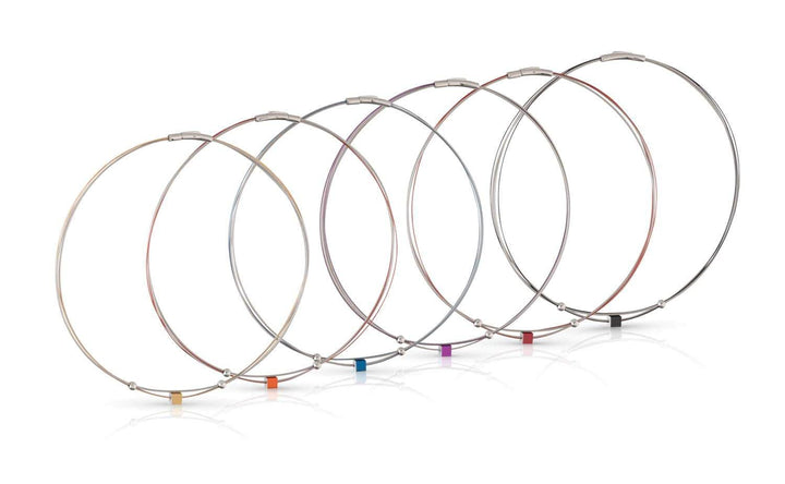 Zeven dunne metalen kabel sleutelhangers van CLIC Creations, genaamd Collier Klein Kubusje Oranje C230O, staan op een rij. Ze hebben elk een kleine kubus met gekleurde labels: geel, oranje, rood, paars, blauw, groen en zwart. De edelstalen bajonet sluiting zorgt voor een veilige pasvorm en glanzende afwerking.