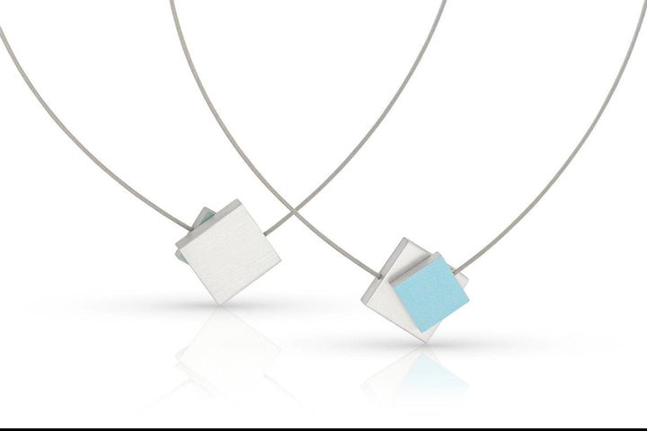 Collier Vierkant met gekleurd vierkant C263B van CLIC Creations bestaat uit twee minimalistische kettingen: één met een blauw vierkant over een wit vierkant, en een andere met een effen wit vierkant. Beide hangers hangen aan dunne zilveren koorden met magnetische sluitingen, prachtig weerspiegeld op een wit oppervlak.