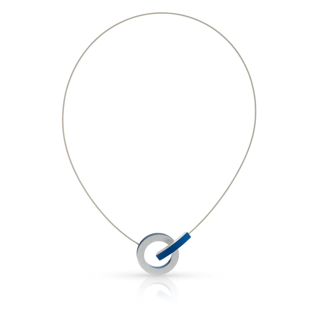 De Collier Ring met gekleurd boogje C273B van CLIC Creations is een minimalistische ketting met een dunne draad en een ronde zilveren hanger met een blauwe accentbalk, geaccentueerd tegen een witte achtergrond. Het bevat een magnetische sluiting en is handgemaakt in Nederland voor een elegante, moderne look.