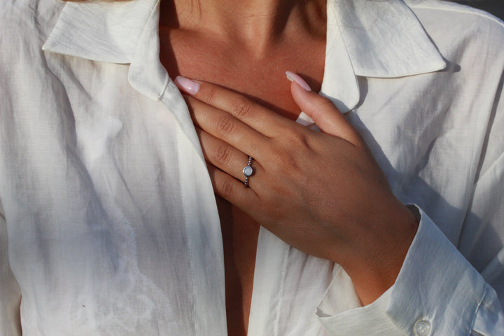 Een persoon met verzorgde nagels, in een wit licht open shirt, raakt zijn borst aan en toont elegant de See You Isabelle | AsRing Zilver. Deze sterling zilveren ring is versierd met een ronde witte steen, die zijn look perfect aanvult.