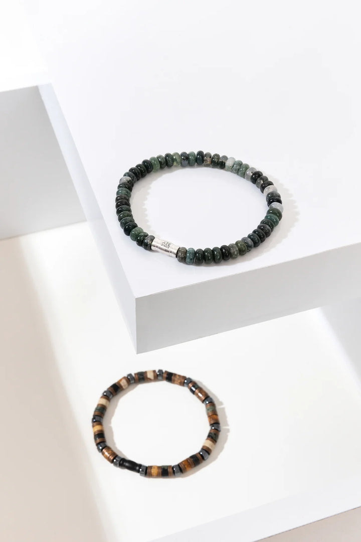 Twee armbanden worden getoond op witte geometrische platforms. De Jade Mountain Oval van AZE Jewels heeft donkergroene kralen met een klein metalen accent, terwijl de Herren-armband afwisselend zwarte en bruine kralen toont.
