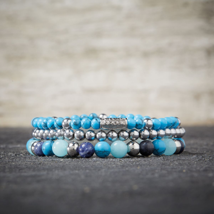 De Rebel & Rose Silver Shine - 4mm Kralen Armband Dames heeft drie gestapelde kralenarmbanden met turquoise, zilveren en blauwe kralen plus een rechthoekige zilveren gegraveerde bedel, afgebeeld op een grijs gestructureerd oppervlak.