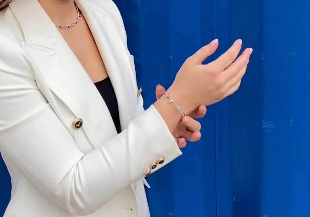 Een persoon in een wit jasje en een zwart topje past de Armband speelse kubusjes zilver A257 van CLIC Creations om zijn pols, tegen een levendige blauwe achtergrond die de elegantie van het tafereel benadrukt.
