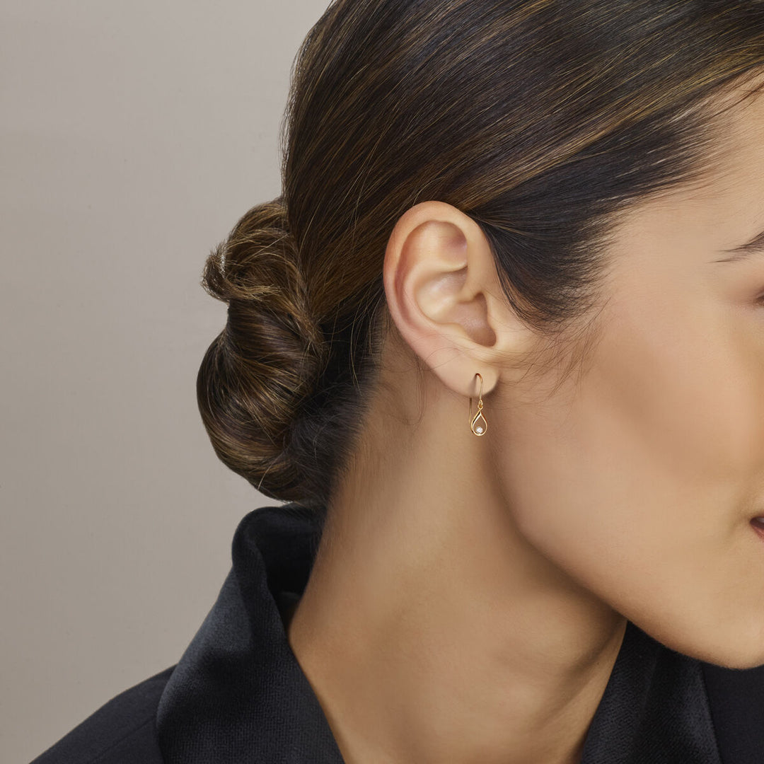 Een vrouw met donker haar in een lage knot draagt een Van der Kooij Collectie oorhangers met Franse haak en zirkonia 23 mm van 14K geelgoud. Ze is gekleed in een zwarte top en de afbeelding benadrukt haar oor en profiel tegen een lichtbeige achtergrond.