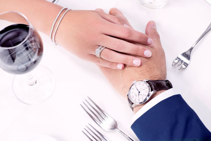 Een close-up van twee handen die elkaar aanraken op een wit tafelkleed: zij draagt de Excellent Jewelry Witgouden Tennisarmband met 63 Briljanten - 3.01 crt Diamant, 14 Karaat en een diamanten ring; hij draagt een horloge. Rode wijn en zilverwerk versterken het elegante tafereel.