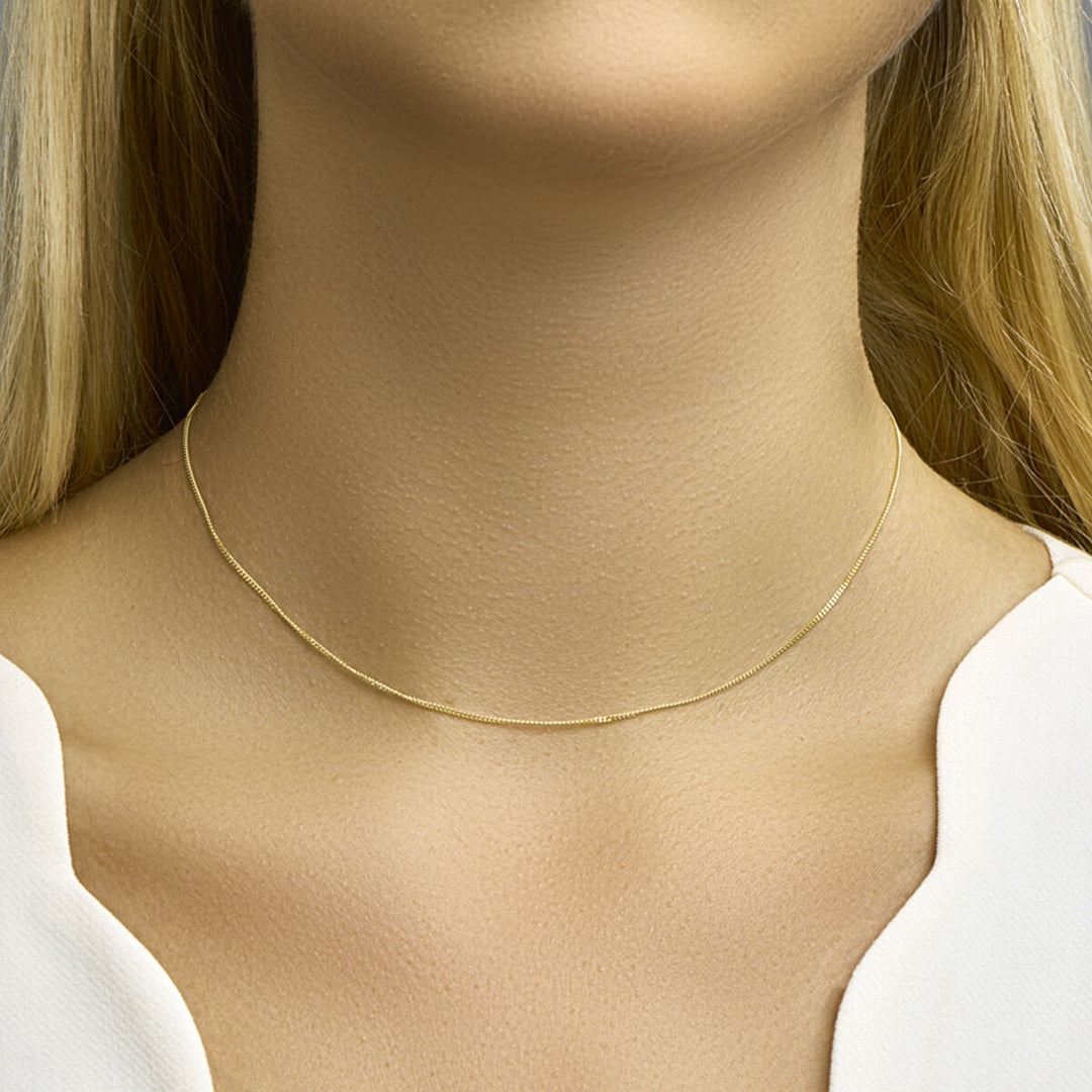 Een close-up van de hals van een vrouw met de Van der Kooij Collectie collier gourmette 1,0 mm 14K geelgoud choker. Ze heeft lang blond haar en is gekleed in een wit topje met een V-vormige halslijn.