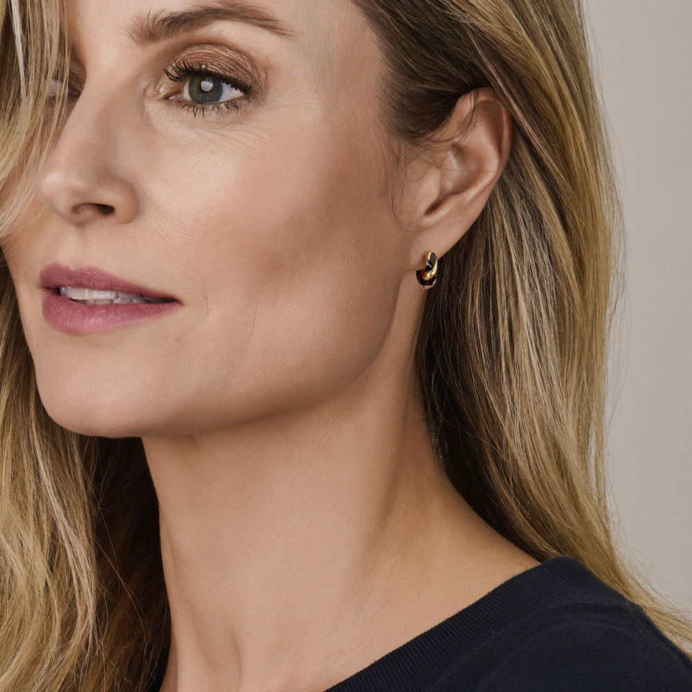 Een vrouw met een lichte huid en blond haar, in een marineblauw topje en Van der Kooij Collectie oorringen zwart gedraaid emaille 1 micron zilver verguld (geel), kijkt iets opzij. Haar make-up is naturel met zachte oogschaduw en roze lippenstift.