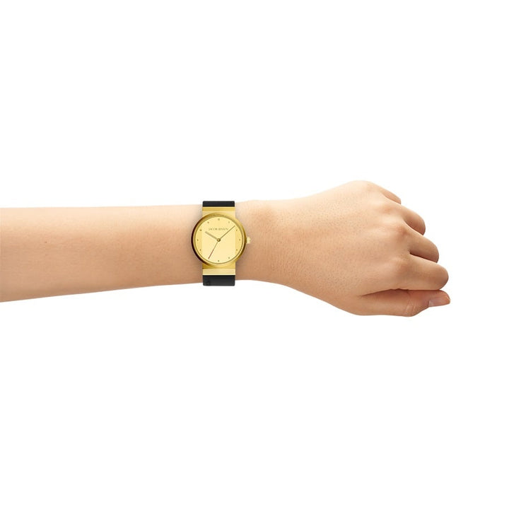 Aan de arm van iemand draagt een Jacob Jensen 745 gouden dameshorloge met een zwarte band, met een minimalistische, ronde wijzerplaat met alleen wijzers op een witte achtergrond die het horloge en de arm elegant in beeld brengt.