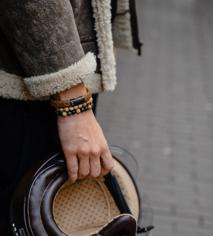 Een persoon in een shearling jas houdt een helm vast, zijn pols versierd met AZE Jewels Ranger Peak 8mm kralenarmband voor mannen. Het wazige bakstenen trottoir op de achtergrond maakt dit stijlvolle ensemble compleet.
