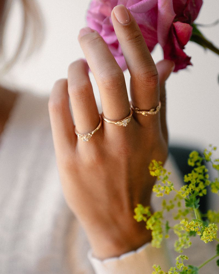 Een hand die drie 14 karaat geelgouden ringen draagt, waaronder de Vintage Driesteens Geelgouden Ring met Ovale Diamant van Excellent Jewelry (0,35 ct, H/SI-1), houdt roze bloemen vast temidden van geelgroene bloesems. De persoon draagt een lichte blouse.