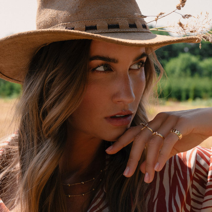 Een vrouw buiten in warm zonlicht, met een breedgerande bruine hoed, gestreept overhemd en de Excellent Jewelry 14 Karaat Geelgouden Damesring (eenvoudig ontwerp, 6 mm ras), poseert nadenkend met onscherp groen achter haar.