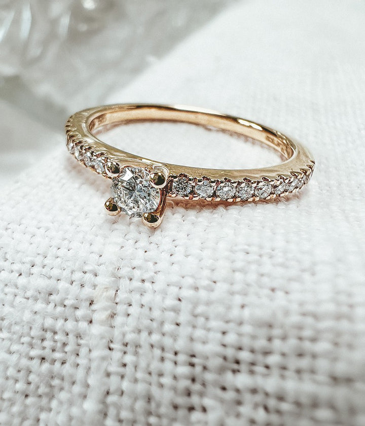 De Schitterende Geelgouden Solitair Ring met Zijstenen van Excellent Jewelry (14 karaat goud, 0.32ct diamant) schittert prachtig op een witte stoftextuur met briljant geslepen middensteen en H/SI-1 diamanten langs de band.
