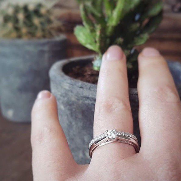 Een hand met de Witgouden Ring met Diamant: Tijdloze Verfijning in 14 Karaat van Excellent Jewelry en een trouwband wordt omhooggehouden voor een cactus, met op de achtergrond een vage plant.