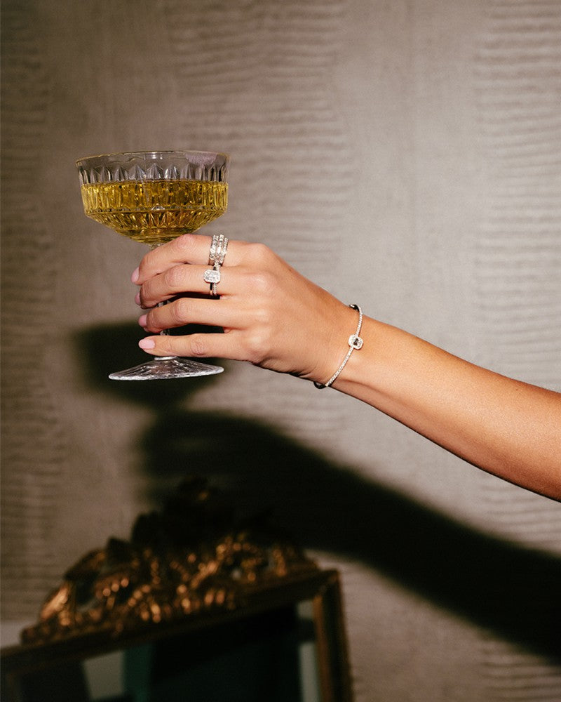 Een gemanicuurde hand die de Excellent Jewelry Elegant White Gold Ring with Brilliant and Baguette Diamonds (0.54 crt) draagt, houdt een vintage glas met gouden drank vast, tegen een gestructureerde achtergrond met een decoratieve spiegel die elegantie toevoegt aan het tafereel.